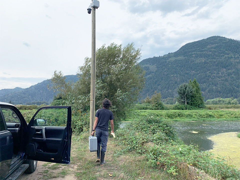 UBC Science Co-op Biology student Elliott Evans sets up water quality monitoring equipment in a green grassy field.