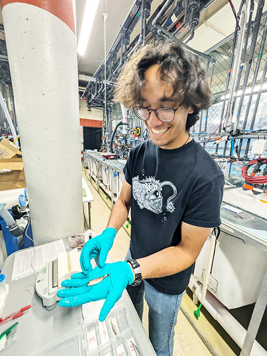 UBC Science Co-op Biology student Elliott Evans holds a tiny fish in his palm in a lab setting.