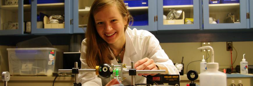 A woman wearing a lab coat smiles at the camera while holding lab equipment.