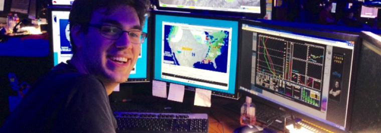 A man smiles at the camera with a computer set up behind him.