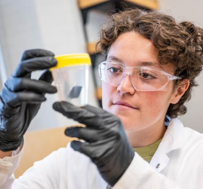 A woman in lab safety gear looks inside a clear container.