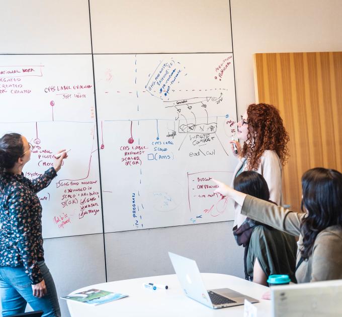 A woman points to complicated diagrams on a whiteboard.