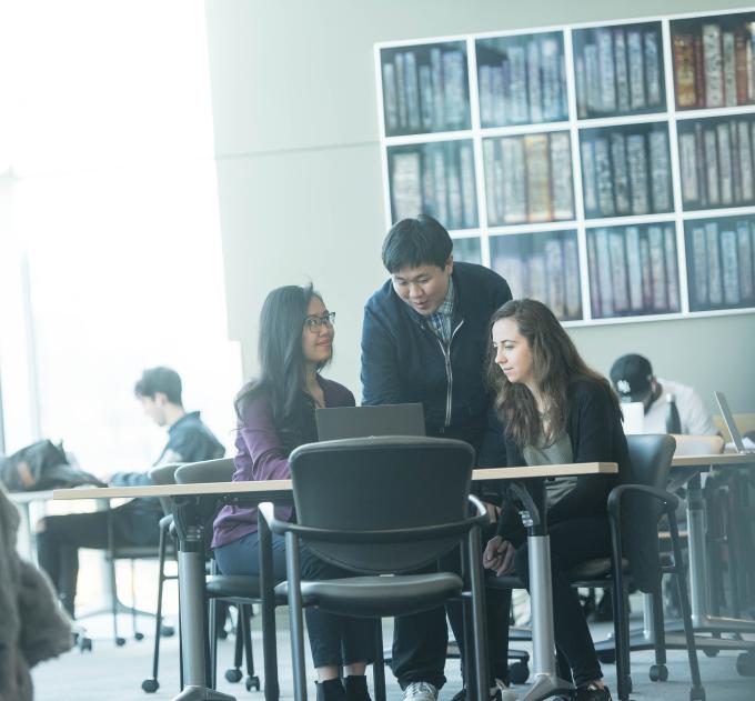 Three students work on a laptop at a communal table.