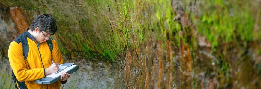 Geological Sciences student examines moss and plant matter on a wall of earth, while making notes.